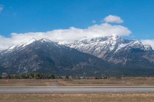 Snow-covered mountains from Grand Teton National Park rise majestically in the background under a clear blue sky The foreground shows part of the Jackson Hole Airport runway with pavement markings