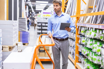 Young man shopping or working in a hardware warehouse standing checking supplies on his tablet.