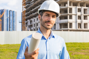 Close-up portrait of a young smiling man architect wearing a helmet with blueprint plans looking at...