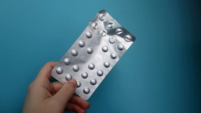 A person holds a silver blister pack of tablets against a blue background. The packaging is partially used, indicating ongoing medication or supplement intake.