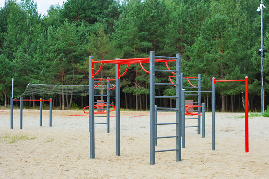 Modern outdoor fitness park with exercise equipment including pull-up bars, parallel bars, and workout stations set on sandy ground surrounded by green forest.