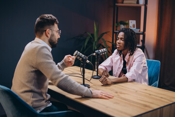 Two male podcasters sitting at a wooden table with microphones, recording a new episode in a...