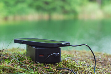 Black portable power bank with smartphone and charging cable on a natural background in nature...