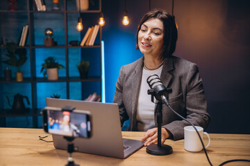 Middle aged female radio host speaking into microphone and recording a podcast and streaming video...