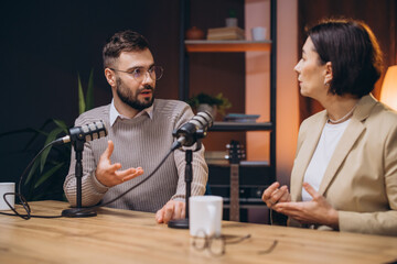 Two podcasters, a man and a woman, are having a conversation and recording a podcast episode in a...
