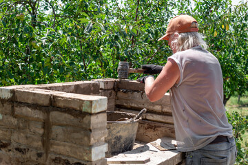 A man builds a brick barbecue structure outdoors, applying mortar and using a rubber mallet, demonstrating the construction process and craftsmanship