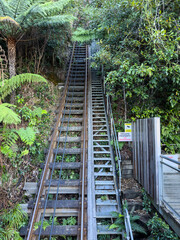 An image of an outdoor funicular railway track and an adjacent metal staircase ascending a steep...