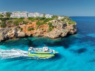 Aerial view of green yacht, sea with clear turquoise water, rocky coast of Cala Mendia in Mallorca, Balearic Islands, Spain. Top drone view of tour boat, houses on the mountain, blue sky at sunset