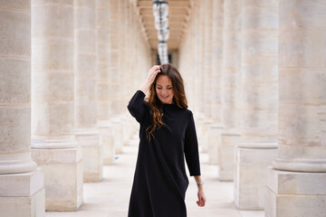 Cheerful young woman in black dress walking confidently through classic architectural colonnade at...