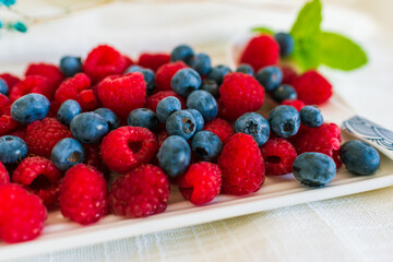 Close-up of fresh raspberries and blueberries on a white plate, decorated with mint leaves and dried flowers. Bright and colorful summer composition.
