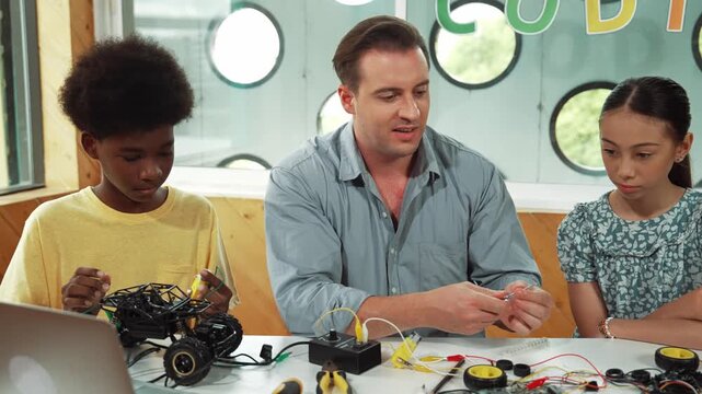 Smart mentor teaching about electronic equipment while fixing car model. Caucasian teacher pointing while explain robotic construction at table with laptop, car model scatter around. Edification