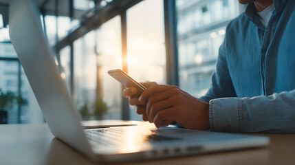 A person working remotely, casual blue shirt, modern office interior with large windows, focused and calm atmosphere, soft morning light, close-up of hands using smartphone and typing on laptop
