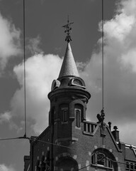 A high, brick tower with a weather vane on the top and some tram electric wires in the grayscale