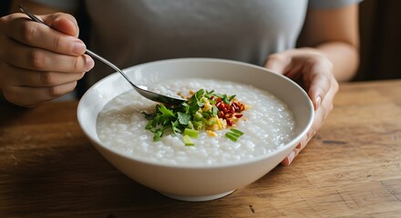 A person enjoying a comforting bowl of traditional Asian rice porridge, or congee, topped with fresh herbs and spices on a wooden table.