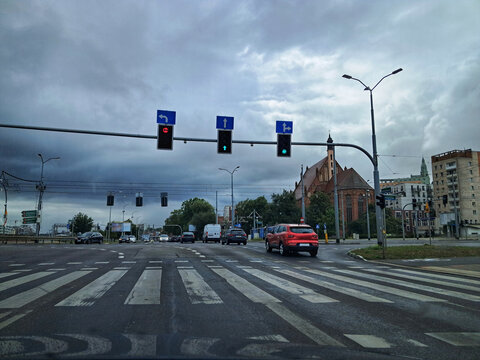 A busy urban intersection in Szczecin, Poland, under a cloudy sky, featuring traffic lights, a pedestrian crossing, and city buildings.