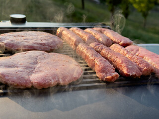 Grilling fresh beef patties and sausages on barbecue, with smoke rising and outdoor greenery in the background, showcasing summer cooking experience
