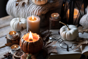Cozy fall table decoration for Thanksgiving, romantic autumn home composition with small pumpkins, candles, wooden knitted sweater, book, dry leaves. Countryside rustic style
