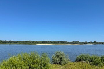 A wide, calm river flows under a clear blue sky, bordered by lush green trees. Concept of: Tranquil nature.