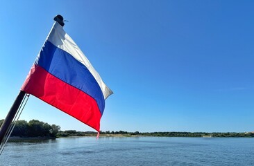 The Russian flag waves against a clear blue sky, with a river landscape in the background.
Concept of: National identity.