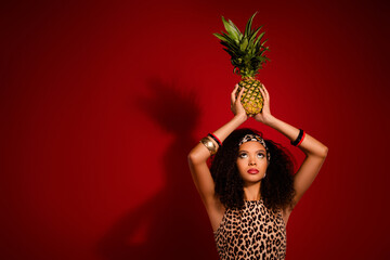 Stylish young woman with afro curly hair holding a pineapple against a vibrant red background in...