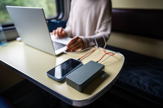 Woman working on laptop, having it and mobile phone connected to modern high-capacity power bank, charging batteries, sits at table in train compartment, selective focus. Remote work while traveling.