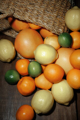 A colorful assortment of citrus fruits, including lemons, limes, oranges, and grapefruits, spills from a woven basket onto a rustic wooden surface. The display highlights a rich variety and texture.