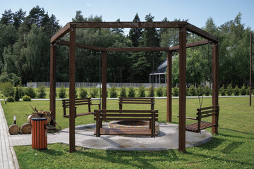 Wooden swing set encircles a fire pit, providing a cozy gathering spot in a green park. Includes three hanging swings supported by a wooden frame on a paved circular base.
