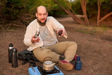 Hispanic male traveler grinding coffee beans in his portable grinder for breakfast outdoors at dawn, in El Palmar National Park, Entre Rios, Argentina.