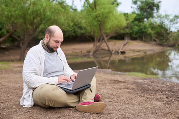 Hispanic man sitting outdoors, in nature, working on his laptop. Concept: digital nomad working remotely while traveling, enjoying a lifestyle of freedom.