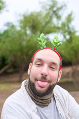 A young Hispanic man wearing a Christmas reindeer antler headband sticks his tongue out at the camera in a playful expression.