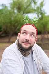 A young Hispanic man wearing a funny Christmas reindeer antler headband looks up with an amused...