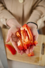 A woman's hands hold pieces of sliced red sweet pepper, top view, a wooden chopping board and a kitchen knife lie in the background
