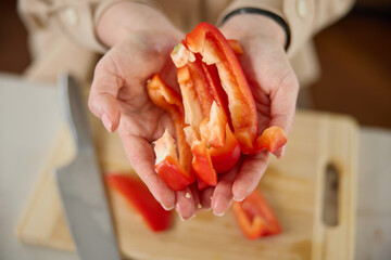 A woman's hands hold pieces of sliced red sweet pepper, top view, a wooden chopping board and a kitchen knife lie in the background