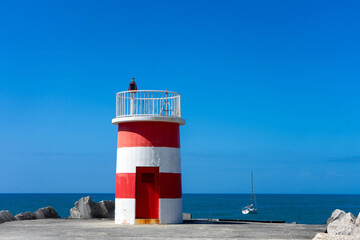 Fototapeta premium Lighthouse overlooking the stunning Nazaré beach, Portugal, with clear blue sky and crashing waves, symbolizing maritime safety and coastal beauty.