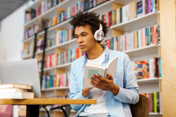 Concentrated Latin student guy in headphones studying online in library with book in front of...