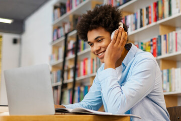Happy African American male student studying in college library, using laptop and wearing wireless...