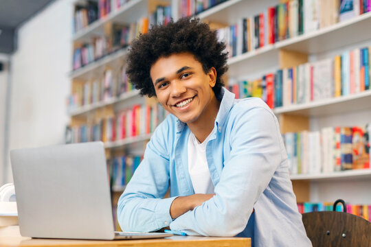 Portrait of happy African American male student sitting in campus library with laptop, preparing to exam, searching information, looking and smiling at camera
