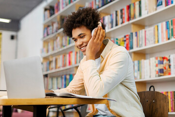 Happy African American male student studying in college library, using laptop and wearing wireless...