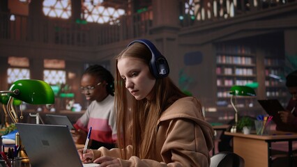 Young student attending to a web lecture with headset in the campus library, takes notes and learns...