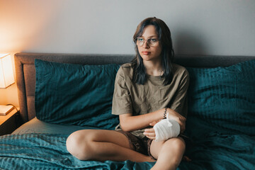 Teenage girl with cast on broken arm sitting on bed at home, recovering and using smartphone at...