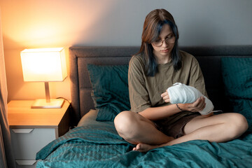 Teenage girl with cast on broken arm sitting on bed at home, recovering and using smartphone at...