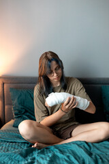 Teenage girl with cast on broken arm sitting on bed at home, recovering and using smartphone at...