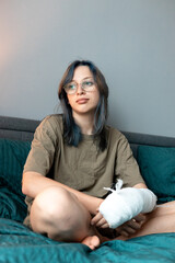 Teenage girl with cast on broken arm sitting on bed at home, recovering and using smartphone at...