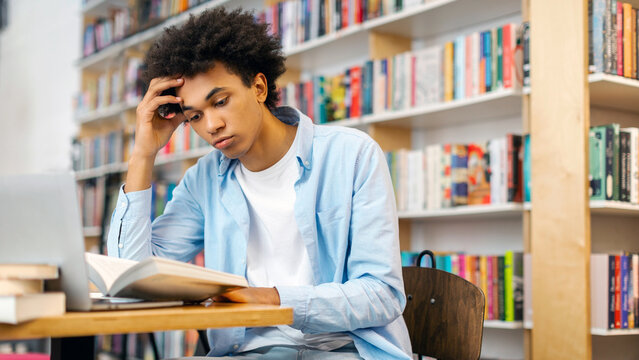 Tired black student guy sitting in campus library at table, reading book, preparing to lecture or exam, free space. Knowledge and learning concept - Powered by Adobe
