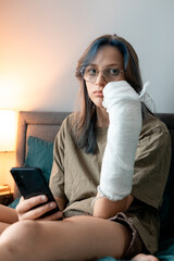 Teenage girl with cast on broken arm sitting on bed at home, recovering and using smartphone at...
