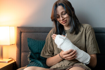 Teenage girl with cast on broken arm sitting on bed at home, recovering and using smartphone at night