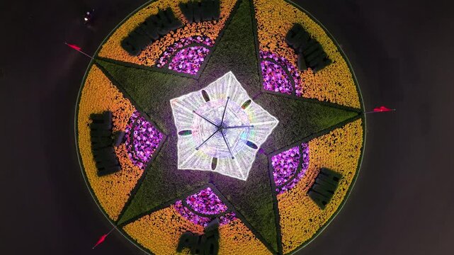 4K top-down drone shot rising vertically from the center of an illuminated floral roundabout at night. Captured in Vietnam, showcasing geometric symmetry and vibrant neon colors.