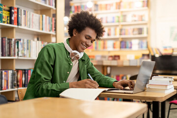 Happy African American student guy using laptop in university library and writing in copybook,...