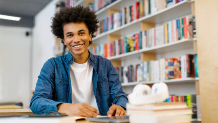 Happy intelligent African American student guy sitting at desk in library and smiling at camera while writing in copybook, free space. Exam preparation concept