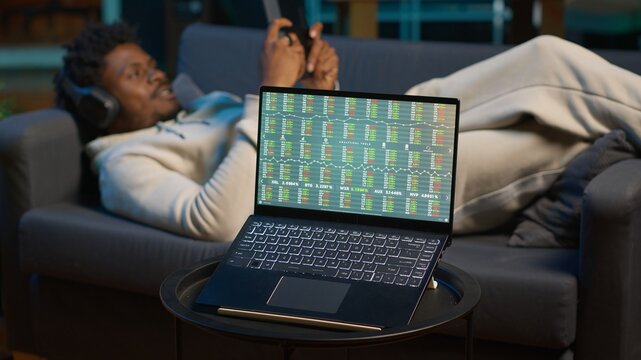 Man enjoying music and reading e books on tablet while waiting for stock market financial figures to get updated on laptop. Person at home next to pc showing trading investment graphs. Camera A.